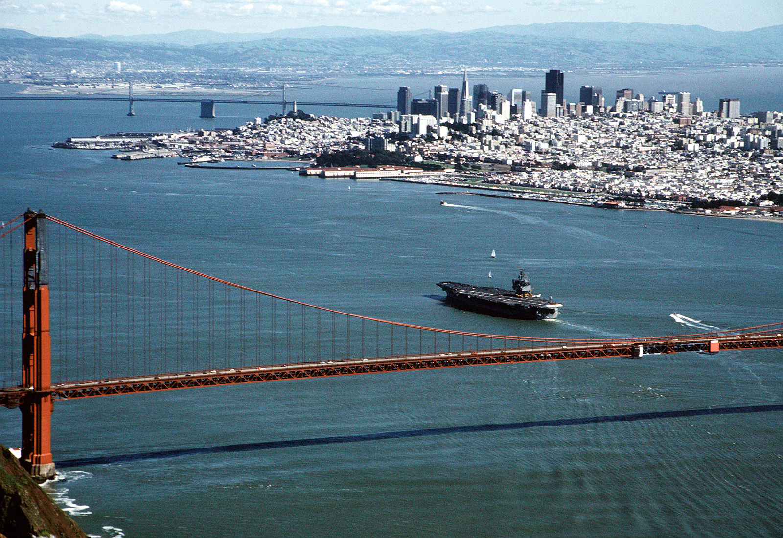 Aerial photo of Golden Gate Bridge and San Francisco skyline with aircraft carrier USS Enterprise having just passed under the bridge entering the harbor